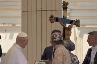 A pope-like figure in white robes receives a framed painting from a woman in a lace dress at a ceremonial gathering, with a crucifix in the background.