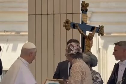 A pope-like figure in white robes receives a framed painting from a woman in a lace dress at a ceremonial gathering, with a crucifix in the background.