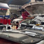 Mechanic in a red shirt leaning over the open hood of a white SUV, inspecting the engine in a busy auto repair shop.