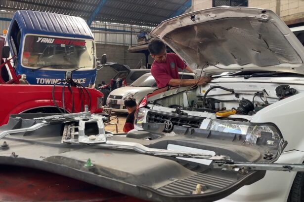 Mechanic in a red shirt leaning over the open hood of a white SUV, inspecting the engine in a busy auto repair shop.