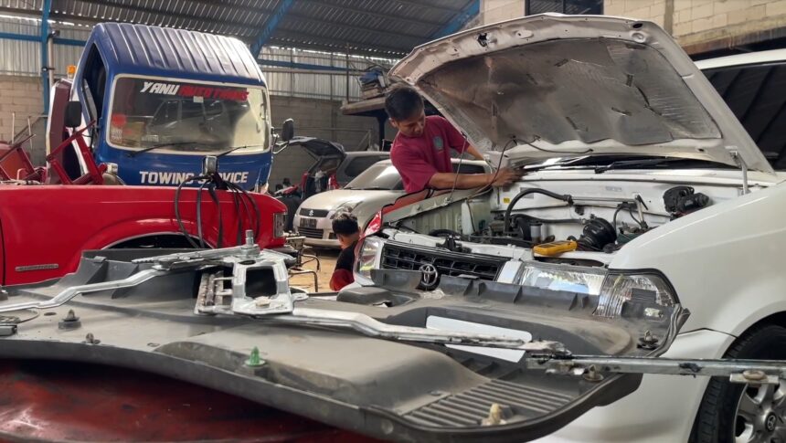 Screenshot_20260619_063203_Drive - Mabur.co Mechanic in a red shirt leaning over the open hood of a white SUV, inspecting the engine in a busy auto repair shop.