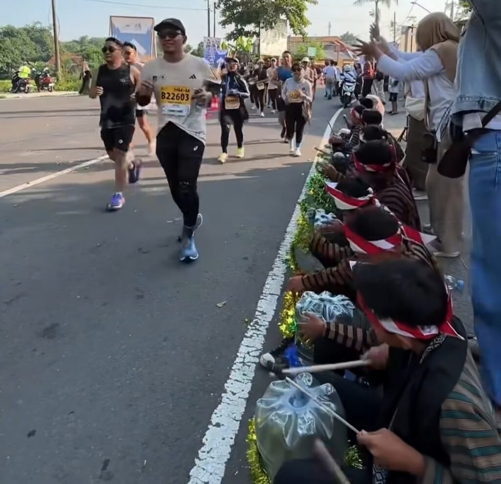 Male runner in a white shirt with bib B22603 runs toward a cheering crowd along a city street curb. Behind him, other runners follow as spectators line the road and clap/cheer.