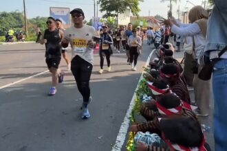 Male runner in a white shirt with bib B22603 runs toward a cheering crowd along a city street curb. Behind him, other runners follow as spectators line the road and clap/cheer.