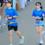 Two runners in matching blue outfits race along a crowd-lined street, bibs visible on their waists.