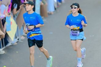 Two runners in matching blue outfits race along a crowd-lined street, bibs visible on their waists.