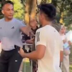 Two men talking in a park: one facing the camera in a gray T-shirt, the other with his back to the camera; trees in the background.