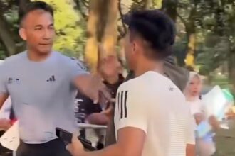 Two men talking in a park: one facing the camera in a gray T-shirt, the other with his back to the camera; trees in the background.