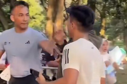 Two men talking in a park: one facing the camera in a gray T-shirt, the other with his back to the camera; trees in the background.