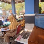Group of people in hijabs seated at a table with a laptop and documents outside, engaging in a service or registration session at a booth.