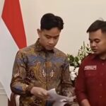 Two men signing papers at a formal ceremony, one wearing a batik shirt and the other in a red uniform, with a national flag in the background.