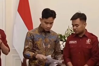 Two men signing papers at a formal ceremony, one wearing a batik shirt and the other in a red uniform, with a national flag in the background.
