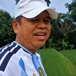 Close-up of a man wearing a white cap and a blue-and-white striped shirt, smiling in a grassy outdoor area with trees in the background.
