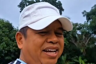Close-up of a man wearing a white cap and a blue-and-white striped shirt, smiling in a grassy outdoor area with trees in the background.