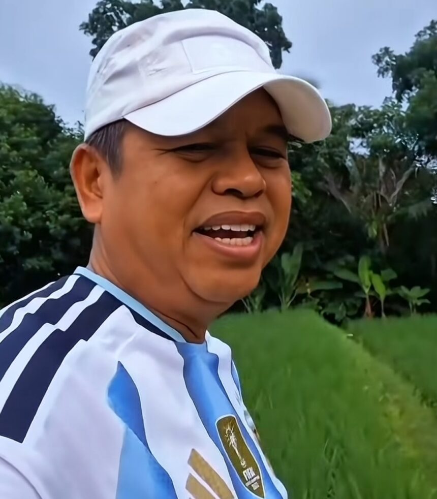 Close-up of a man wearing a white cap and a blue-and-white striped shirt, smiling in a grassy outdoor area with trees in the background.