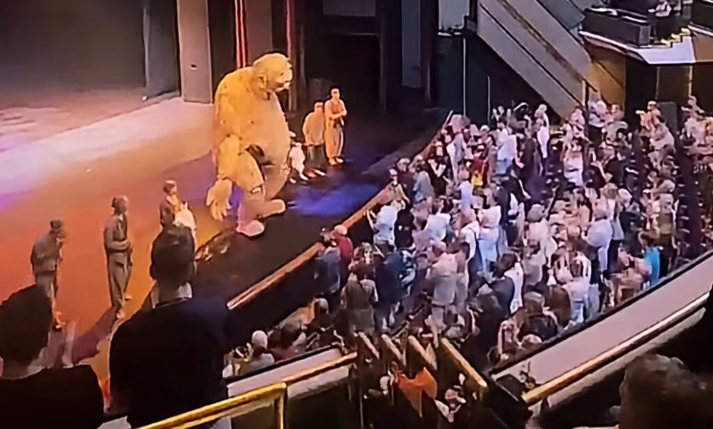 Screenshot_20260623_223516_Instagram - Mabur.co Large yellow bear mascot on a stage beside performers, facing a packed audience in a theater.