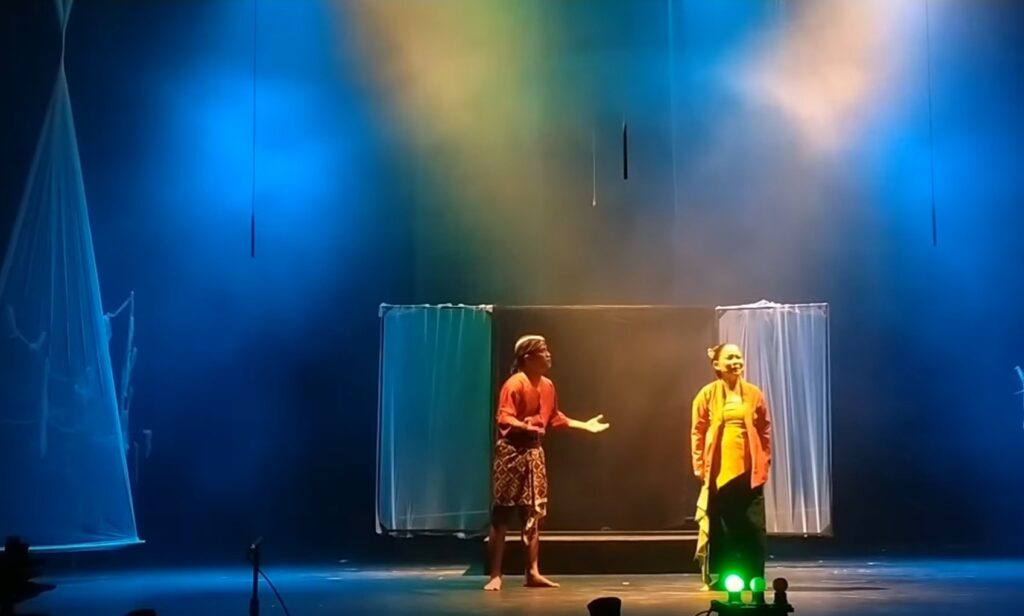 Two performers on a dimly lit stage with blue lighting and hanging fabric panels. One is speaking while the other smiles.