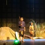 Three performers in period costumes on a stage; one man holds a large prop sword while two others stand nearby amid a camp-like set with grasses and a tent backdrop.