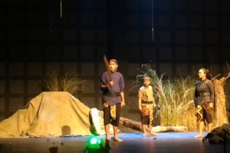Three performers in period costumes on a stage; one man holds a large prop sword while two others stand nearby amid a camp-like set with grasses and a tent backdrop.