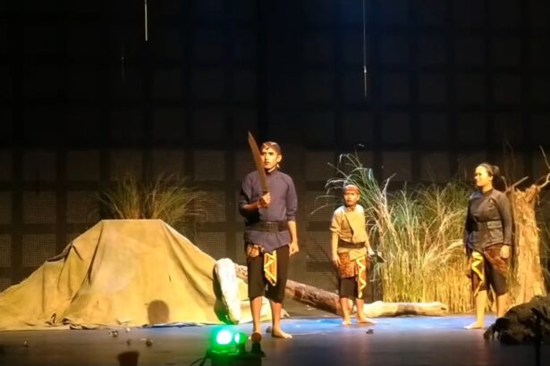 Three performers in period costumes on a stage; one man holds a large prop sword while two others stand nearby amid a camp-like set with grasses and a tent backdrop.
