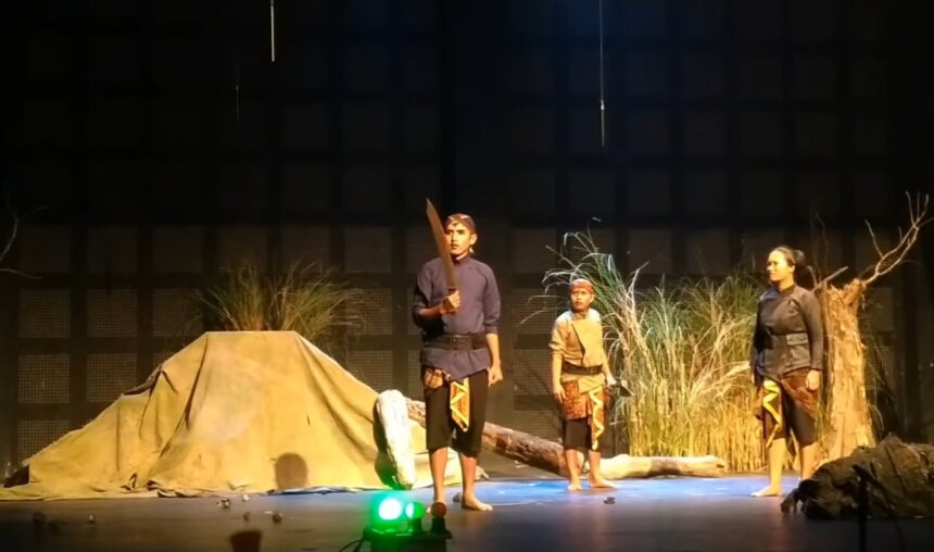Three performers in period costumes on a stage; one man holds a large prop sword while two others stand nearby amid a camp-like set with grasses and a tent backdrop.