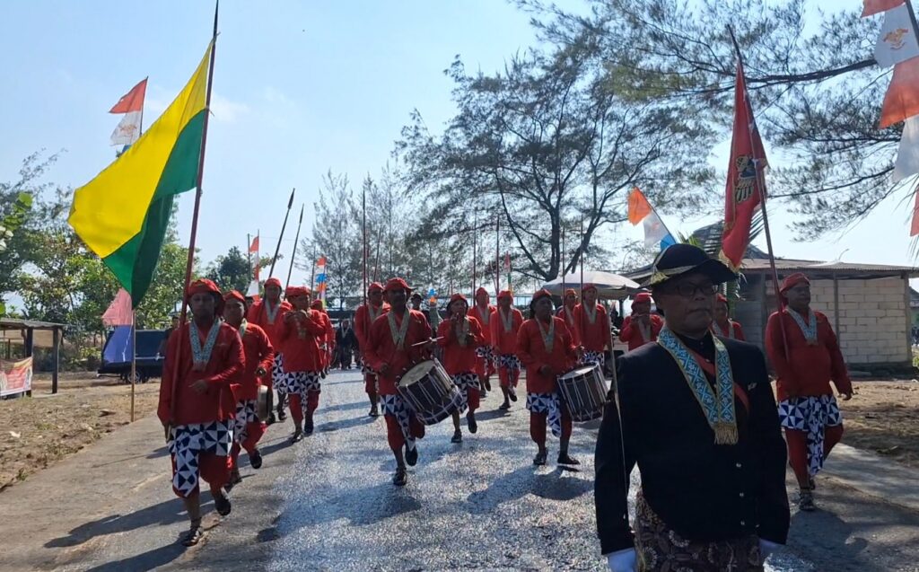 Group of performers in red outfits marching with drums in a flag parade on a sunny day, with colorful flags overhead and a man in a dark suit and hat in the foreground.