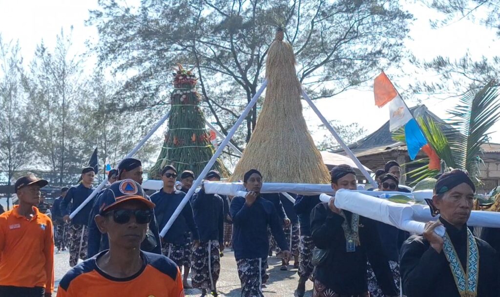 People in matching dark blue outfits participate in a ceremonial parade, carrying a long white pole over their shoulders in front of a tall straw centerpiece and colorful flags.