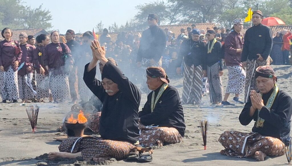 Balinese ceremony on the beach with priests in black, chanting and raising hands over a small fire while a crowd watches in traditional wrap sarongs and batik outfits.