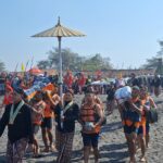 Procession of people in bright red and patterned attire marching along a sandy beach during a cultural festival, with flags and banners in the background.