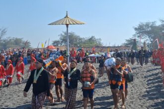 Procession of people in bright red and patterned attire marching along a sandy beach during a cultural festival, with flags and banners in the background.