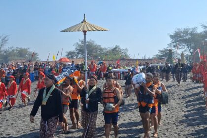 Procession of people in bright red and patterned attire marching along a sandy beach during a cultural festival, with flags and banners in the background.