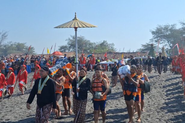 Procession of people in bright red and patterned attire marching along a sandy beach during a cultural festival, with flags and banners in the background.