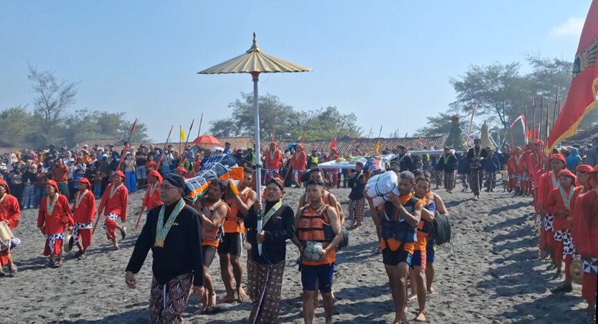 Procession of people in bright red and patterned attire marching along a sandy beach during a cultural festival, with flags and banners in the background.