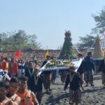 Groups of people in bright traditional clothing carry a tall, decorated ceremonial structure along a sandy beach during a festival, with colorful flags in the background.