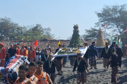 Groups of people in bright traditional clothing carry a tall, decorated ceremonial structure along a sandy beach during a festival, with colorful flags in the background.