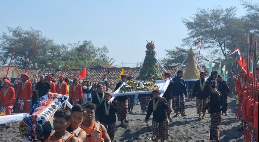 Groups of people in bright traditional clothing carry a tall, decorated ceremonial structure along a sandy beach during a festival, with colorful flags in the background.