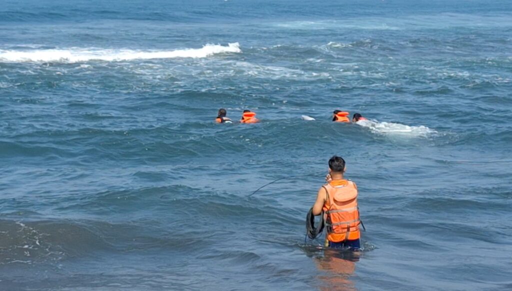 A group of people in orange life jackets afloat in the ocean, with a supervising person in a bright orange vest standing in the water nearby.