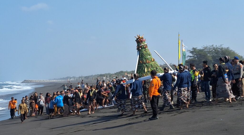 Crowd gathers on a dark-sand beach for a traditional ceremony as a tall decorated bamboo structure is carried forward.