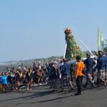Crowd gathers on a dark-sand beach for a traditional ceremony as a tall decorated bamboo structure is carried forward.