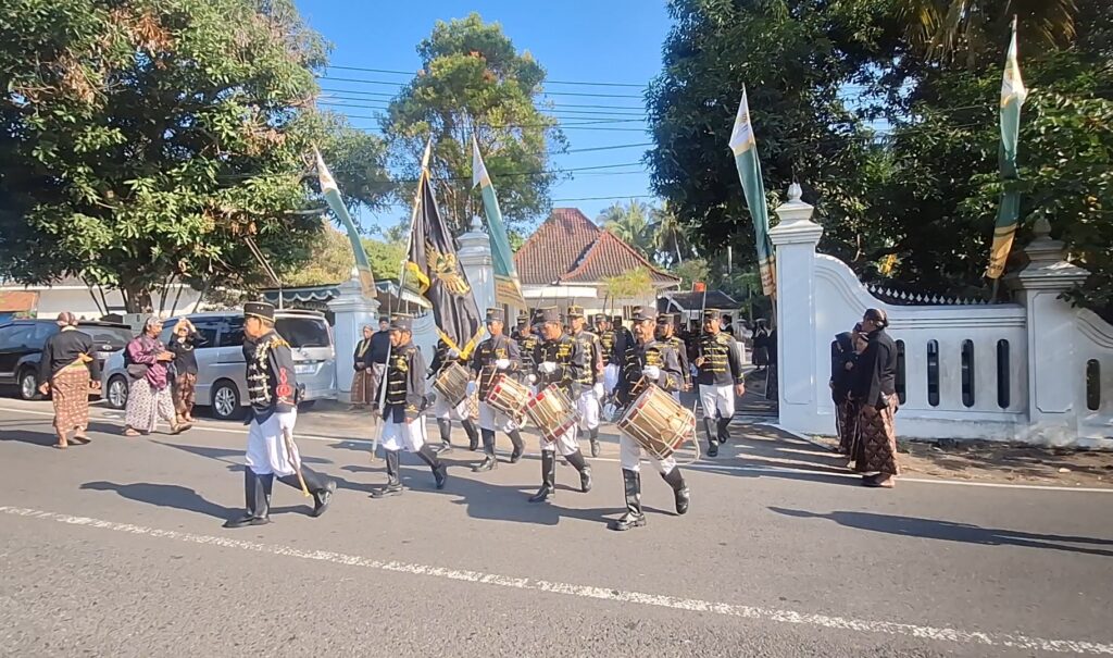 Group of drummers in ceremonial black-and-gold uniforms marching with flags during a sunny street parade, spectators watching from the sidewalk.