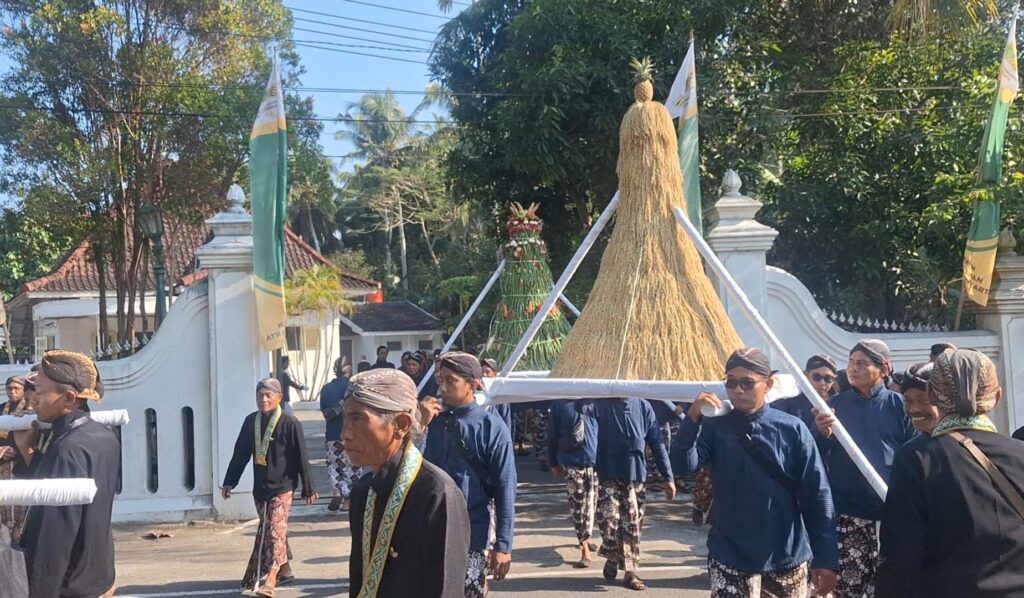 Ceremonial procession with men in traditional clothing carrying a tall straw sculpture on poles, near a white gate and green banners.
