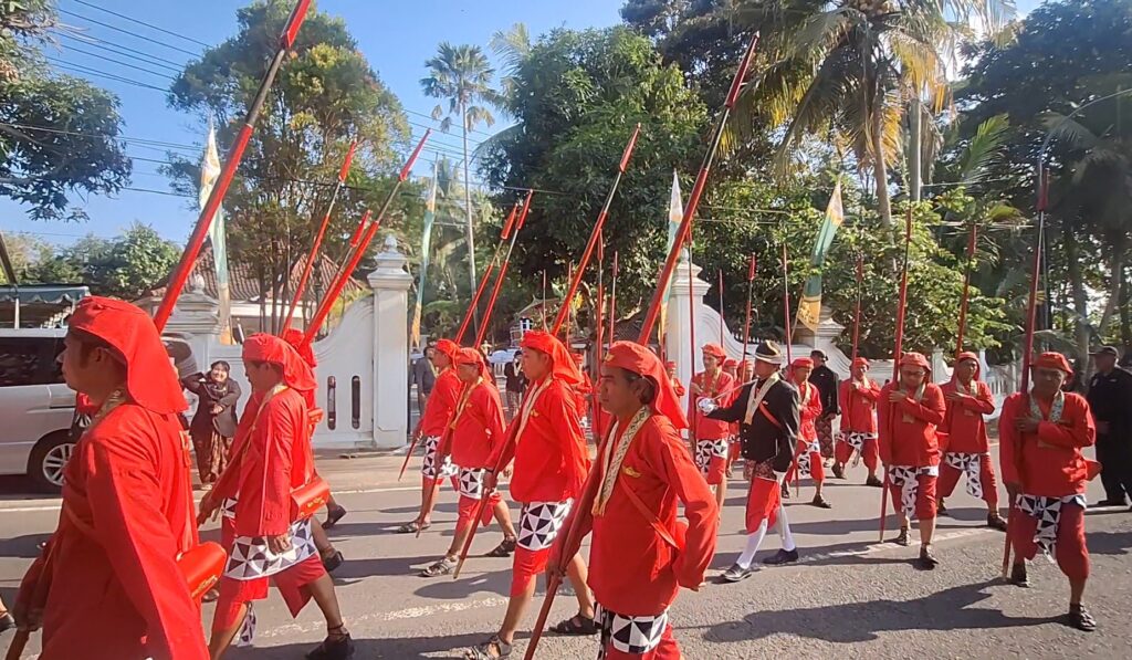 Group of red-clad parade participants marching with long red spears along a street, palm trees in the background.