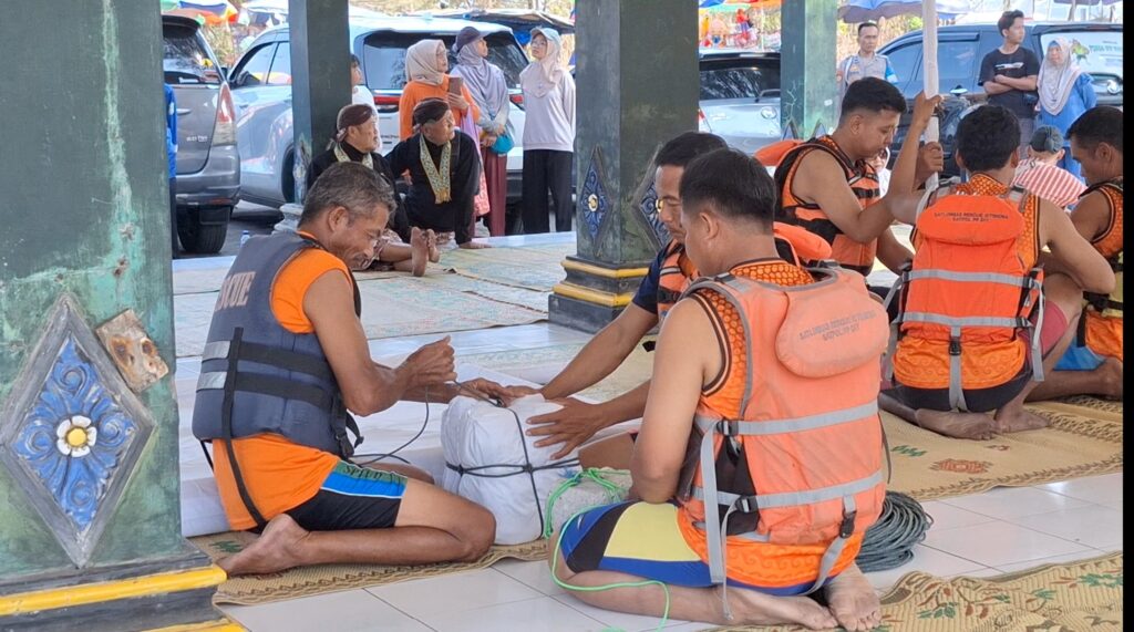 Rescue workers in orange life jackets kneel on mats around a person on a white sheet during a training drill under a shaded area, with onlookers nearby.
