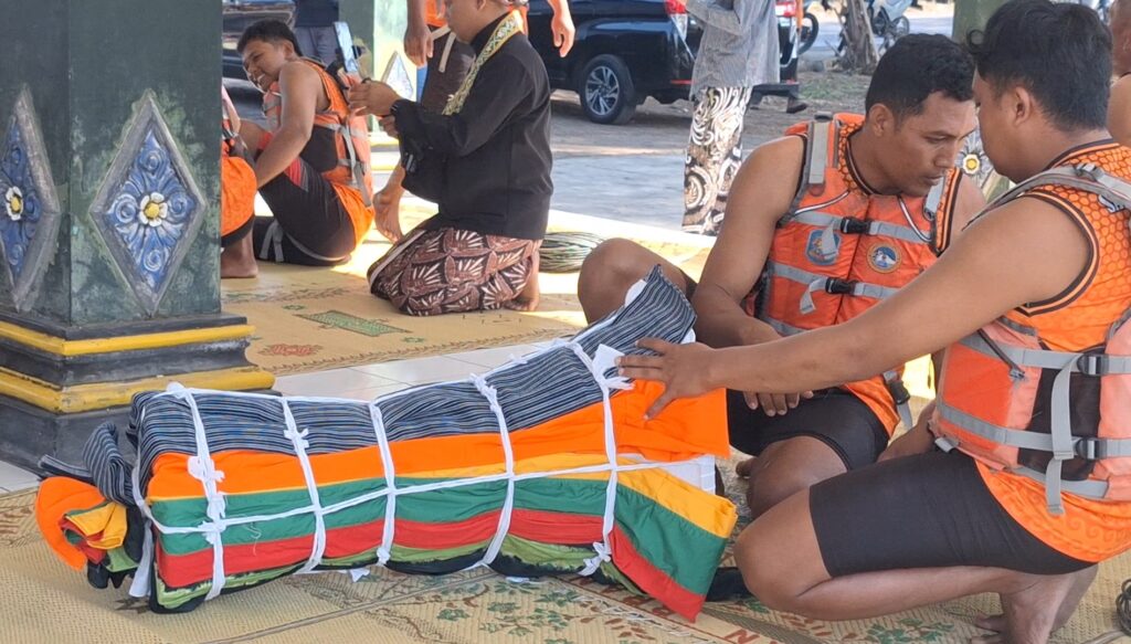 Rescue workers in orange life jackets kneel around a body wrapped in a colorful, flag-like bundle on a mat, preparing for transfer.