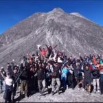 Group of hikers on a rocky mountain summit, arms raised in celebration, with a tall peak and clear blue sky behind them.
