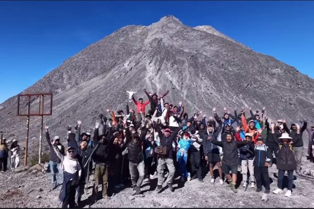 Group of hikers on a rocky mountain summit, arms raised in celebration, with a tall peak and clear blue sky behind them.