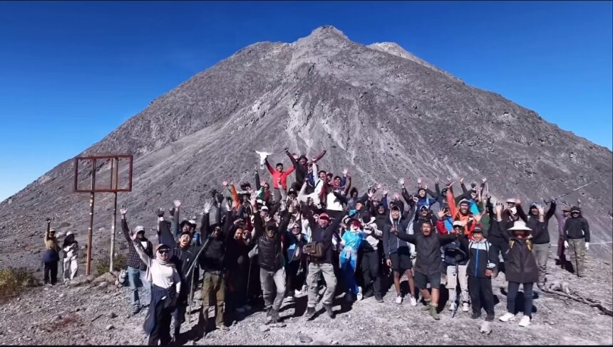Group of hikers on a rocky mountain summit, arms raised in celebration, with a tall peak and clear blue sky behind them.