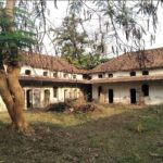 Abandoned white building with arched doorways around a dirt courtyard, a large tree in the foreground and a pile of branches in the center.