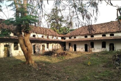 Abandoned white building with arched doorways around a dirt courtyard, a large tree in the foreground and a pile of branches in the center.