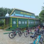 Green mosque with yellow trim and decorative lattice walls, named Masjid Jami MTBFI, and a row of bicycles parked outside with people nearby.