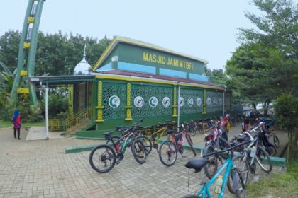 Green mosque with yellow trim and decorative lattice walls, named Masjid Jami MTBFI, and a row of bicycles parked outside with people nearby.